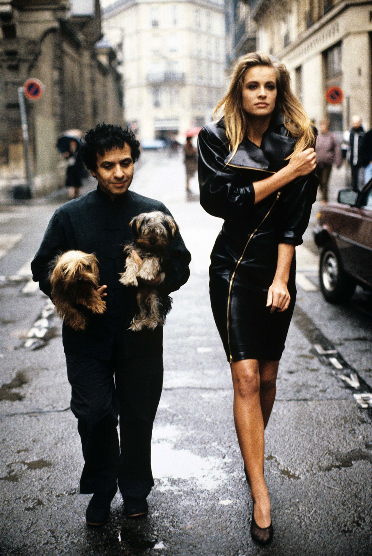 Fashion designer Azzedine Alaia holding his two Yorkshire terriers, Patapouf and Wabo, walking in Paris street with model Frederique who wears one of his creations, a black leather zippered dress, 1986. Photo by Arthur Elgort.